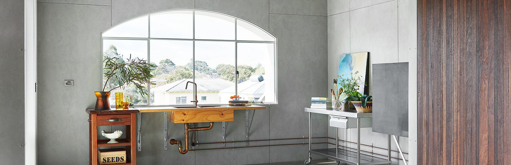 A bright room with a large arched window overlooking houses and trees. Inside, there's a wooden sink with exposed pipes, a wooden cabinet with dishes and a "Seeds" box, and a metal cart with plants and kitchenware. Walls are gray, and the floor is concrete.