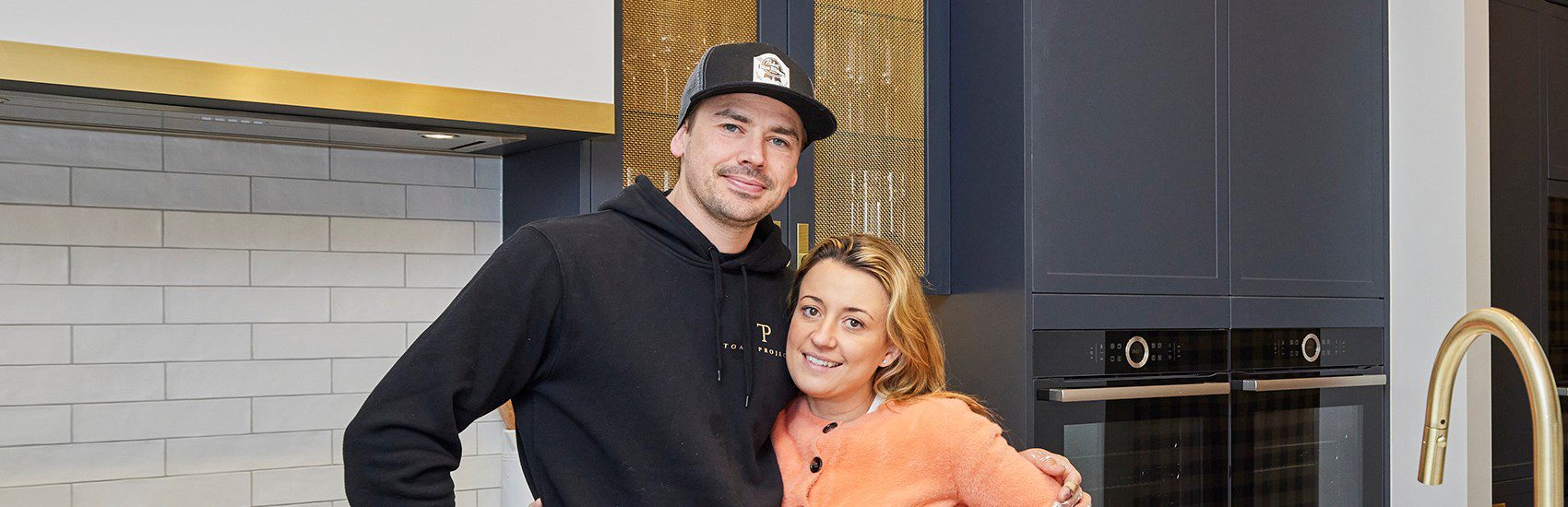 A man wearing a black hoodie and cap embraces a woman in an orange sweater in a modern kitchen. The kitchen features a dark blue cabinet, gold faucet, and a double oven. Both are smiling at the camera.