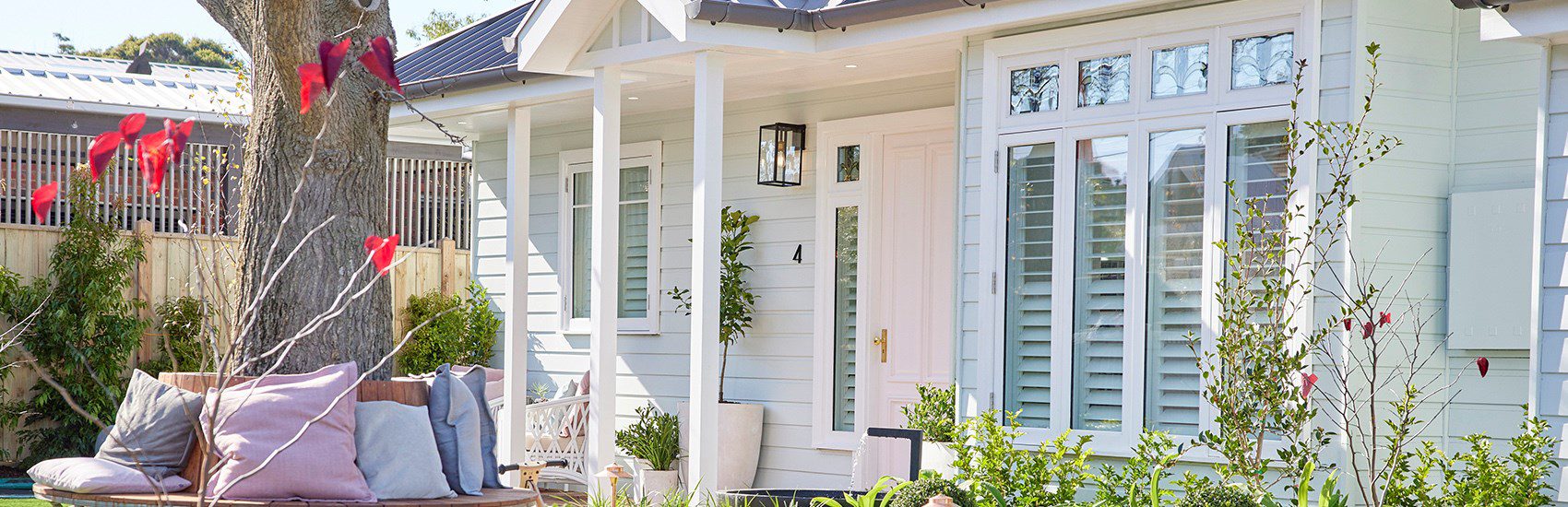 A charming white house features large windows with shutters and a welcoming front porch. The yard is adorned with colorful flowers and a comfortable outdoor seating area with cushions under a tree. The sun casts a soft light on the serene, well-landscaped garden.