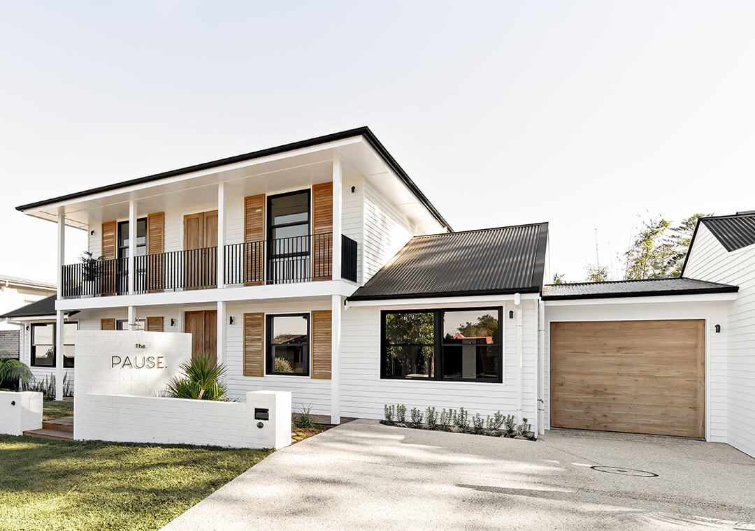 A modern two-story white house with black trim and a wooden garage door. The front yard features a small grass lawn and a driveway. A sign reads "PAUSE" at the front of the property near a white mailbox. The house has a covered balcony with wooden shutters.