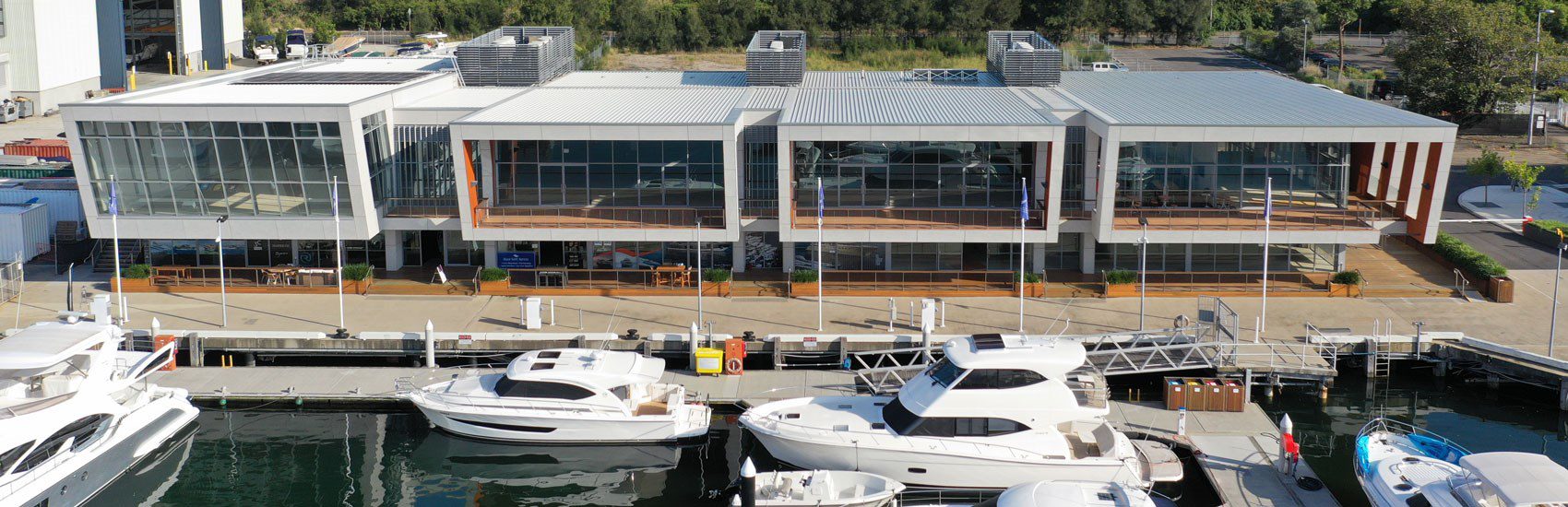 A modern, two-story marina complex with large glass windows overlooks a dock where several luxury boats and yachts are moored. The building features a sleek design with metal and wood accents. Surrounding the marina are green trees and additional industrial buildings.