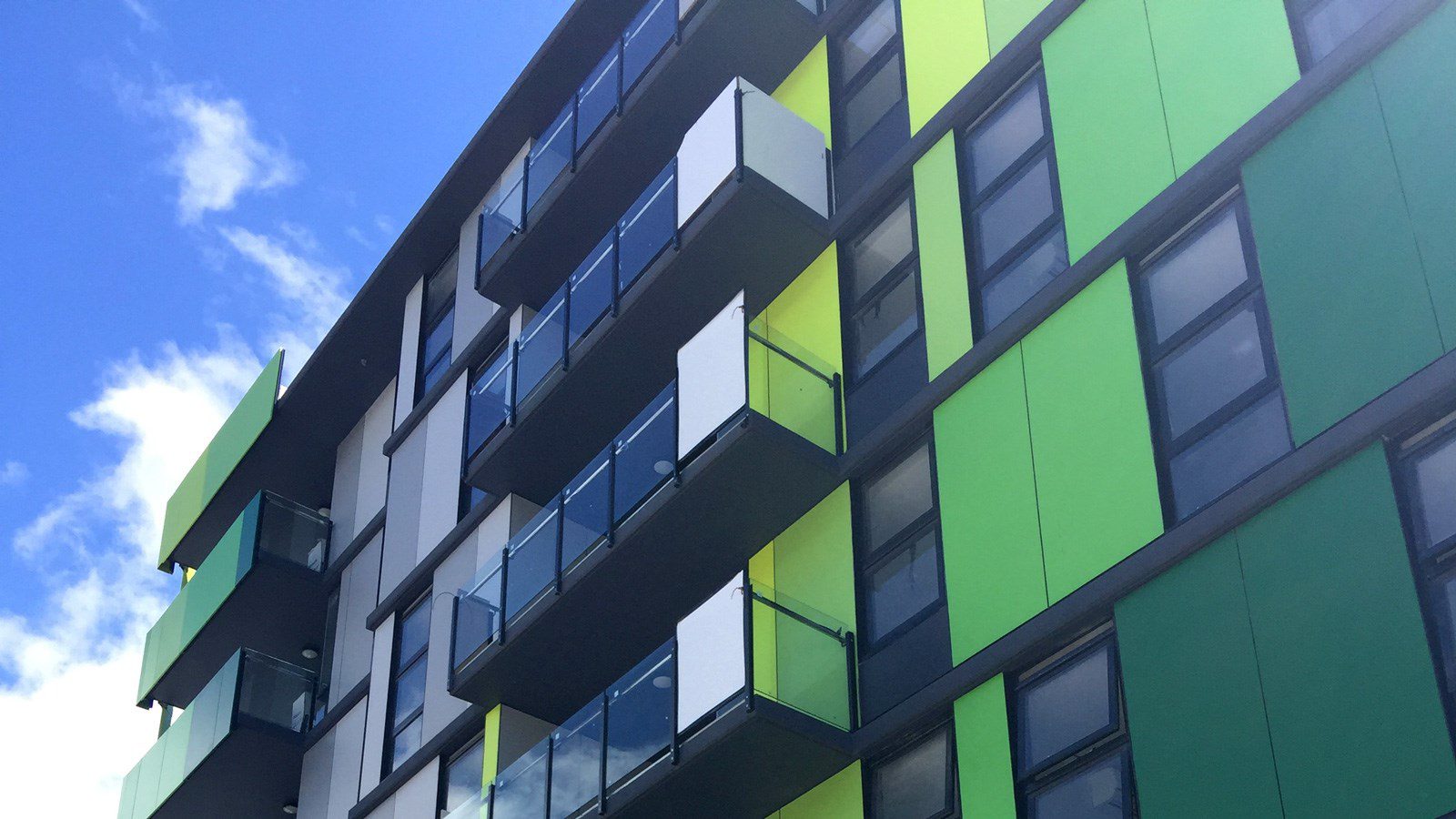 A modern apartment building with vibrant green, yellow, and gray panels under a clear blue sky. The building features multiple balconies with glass railings.