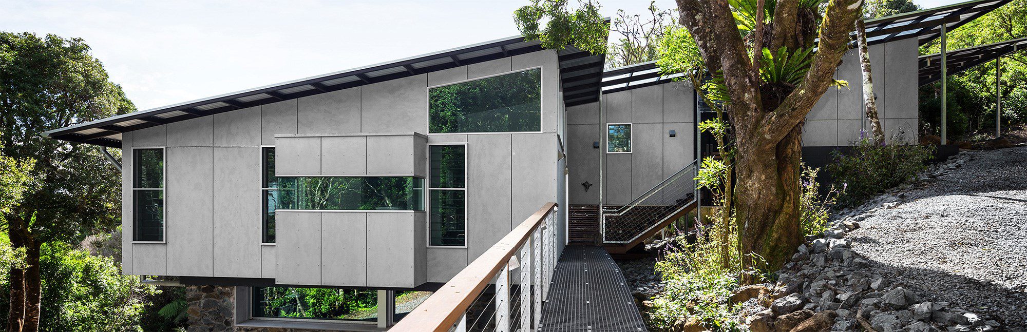 A modern house with a unique, angular design is set on a sloped, rocky terrain amidst dense greenery. It features large windows and a light gray facade. A metal walkway leads to the entrance, and trees surround the structure.