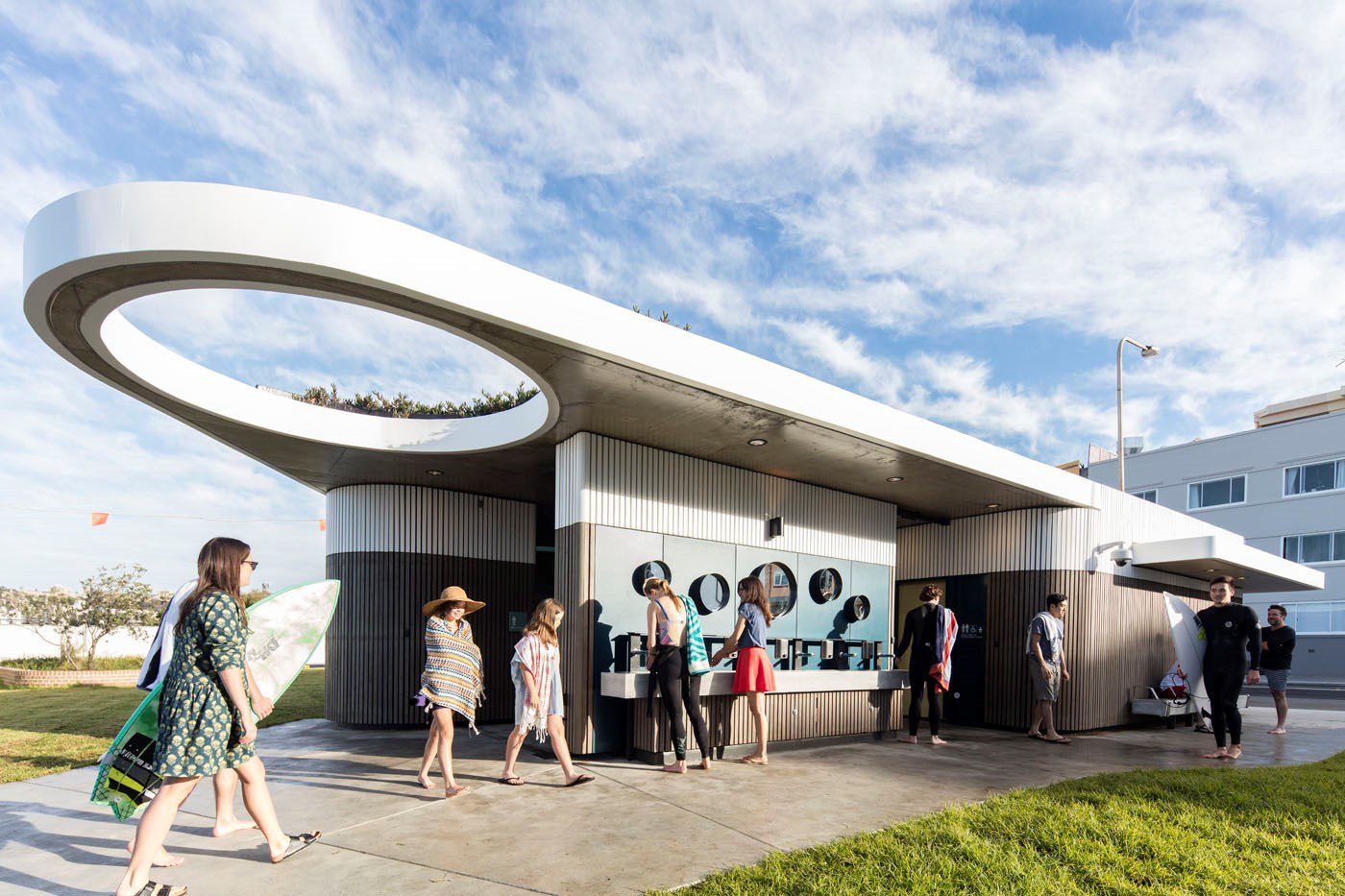 A group of people, some in swimwear, gather around a modern, cylindrical kiosk at a beach. The building has circular windows, sleek metallic accents, and a unique overhang structure. A person with a surfboard walks by, and a clear sky is visible overhead.