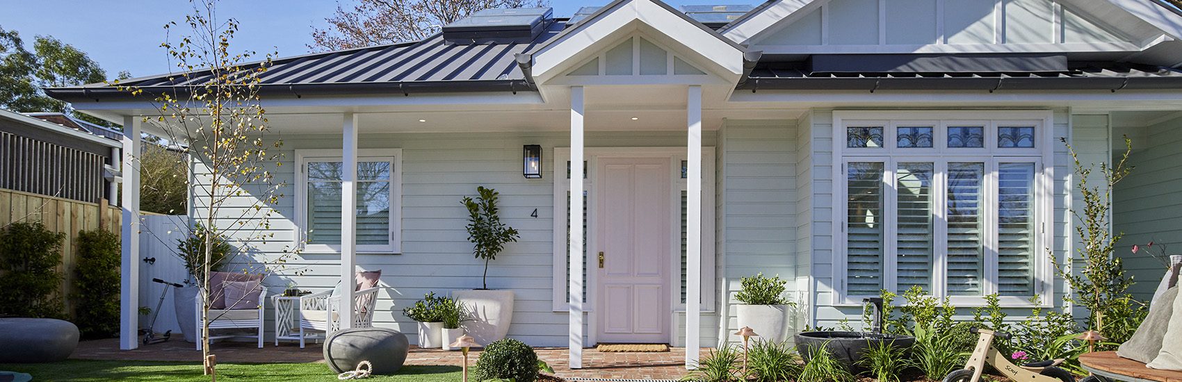 A charming house with light blue exterior walls, a dark metal roof, and a white front door with sidelights. A porch with white pillars and a rocking chair is decorated with plants and garden ornaments. The yard features a lawn and potted plants.