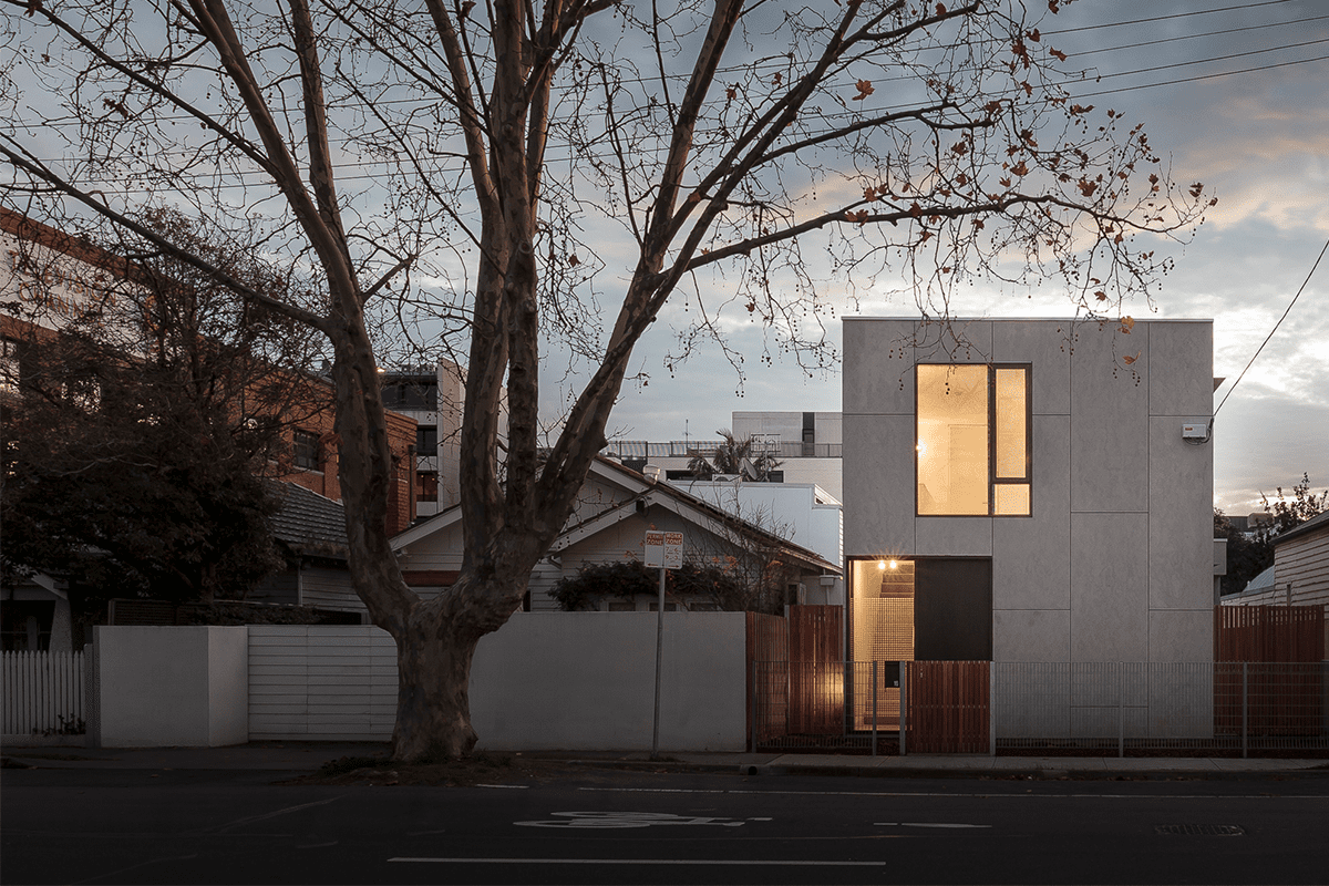 A modern, two-story house with a minimalist design is illuminated warmly from within at dusk. The house features large glass windows and a sleek exterior. A bare tree stands prominently in the foreground, and neighboring homes are visible to the left.