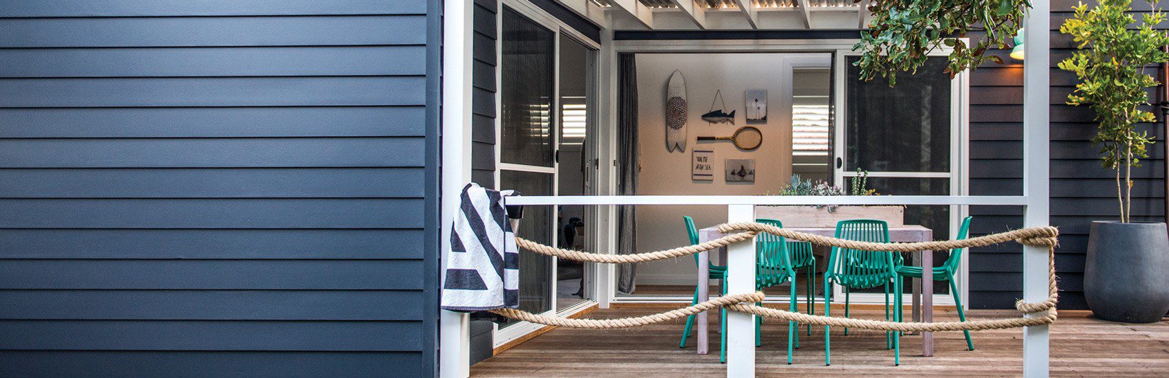 A cozy outdoor patio with blue wooden siding, a white railing adorned with rope, and a dining table surrounded by green chairs. The area is decorated with wall art of surfboards and nautical elements. A towel hangs over the railing, and a potted plant adds greenery.