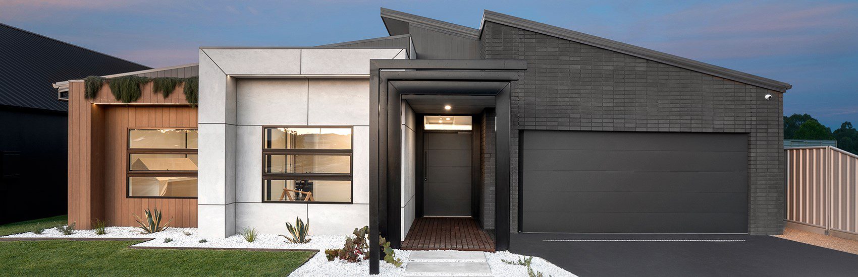 A modern, single-story house with a mix of stone, wood, and dark-colored facade elements. It features a geometric roof design, a large double garage, and a well-maintained front yard with white gravel and plants. The entrance is highlighted by a narrow, covered walkway.