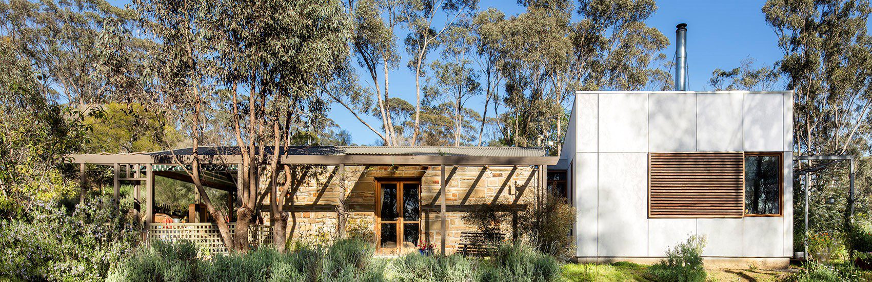 A house with a rustic and modern blend featuring a stone and wood section on the left and a white, minimalist section on the right. It is surrounded by large trees and lush vegetation, with a clear blue sky in the background.