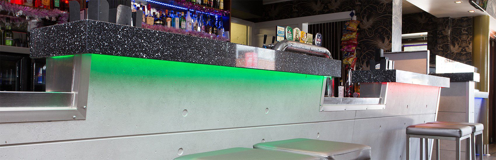 A modern bar with a sleek, speckled black countertop illuminated by vibrant underlighting in green and red hues. Various bottle displays and tap handles are visible in the background, and shiny metal bar stools are positioned under the counter.