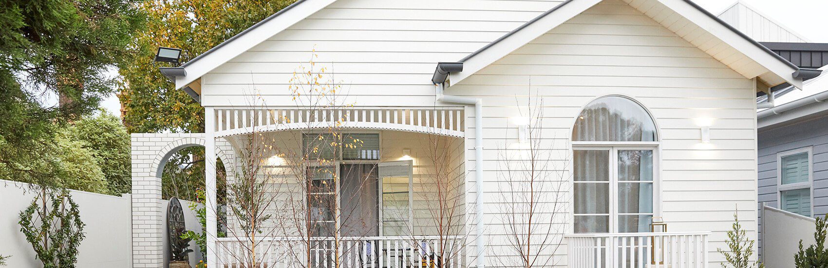 A white, single-story house with a gable roof and a veranda. The veranda has a decorative wooden railing and an arched entryway. Leafless trees and shrubs are in the front yard, and warm lights illuminate the porch area.