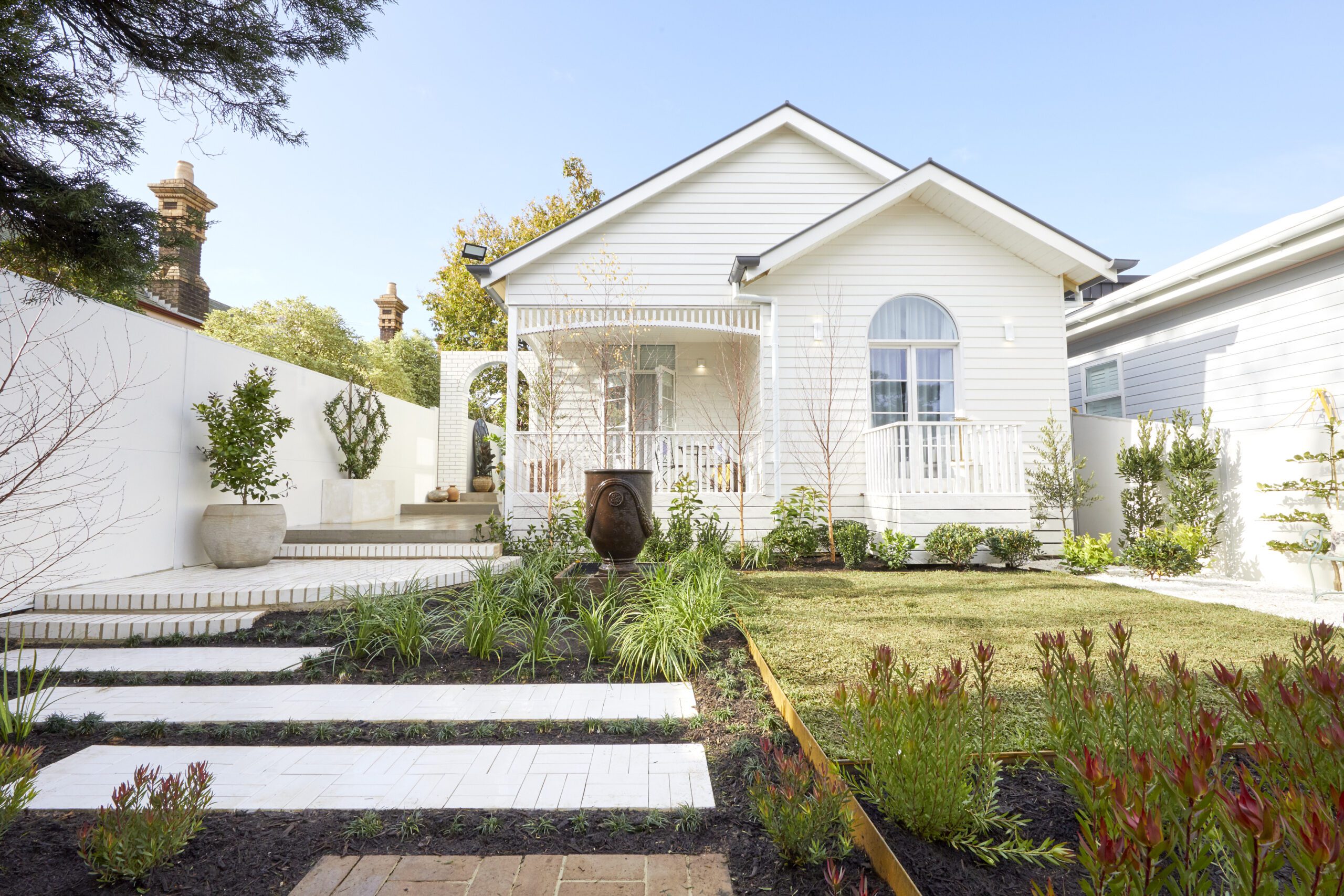 A white house with a front porch, arched window, and neat landscaping featuring green grass, shrubs, and stone pathways leading through a tidy garden under a clear blue sky.