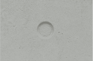 A close-up of a textured, light gray surface featuring a circular indentation in the center. The surface appears to be made of concrete or a similar material with small pores and a slightly rough texture.