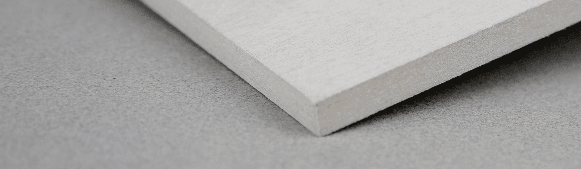 Close-up image of the edge of a rectangular, white, fibrous material sheet placed on a light gray surface. The sheet's texture and thickness are evident, showing a clean, cut edge and smooth surface.