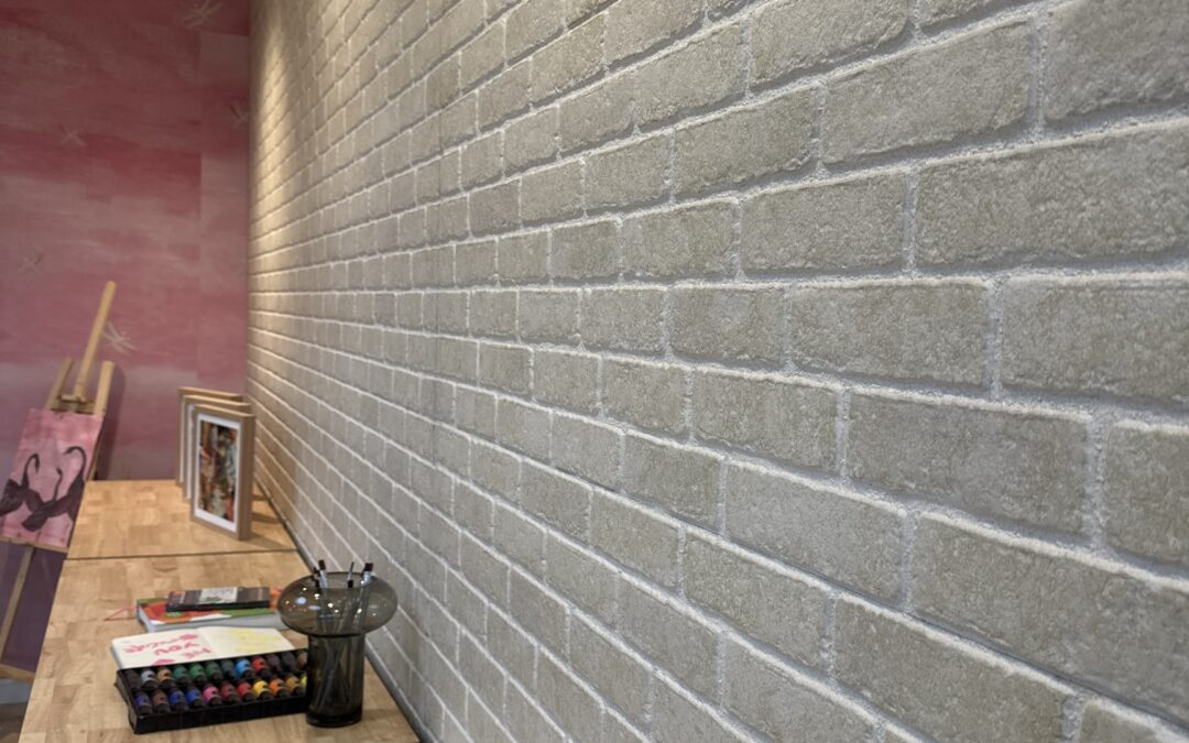 A wooden desk runs alongside a light-colored brick wall. Art supplies, colored pencils, a framed picture, and a painting on an easel are arranged on the desk. The far wall is painted pink.