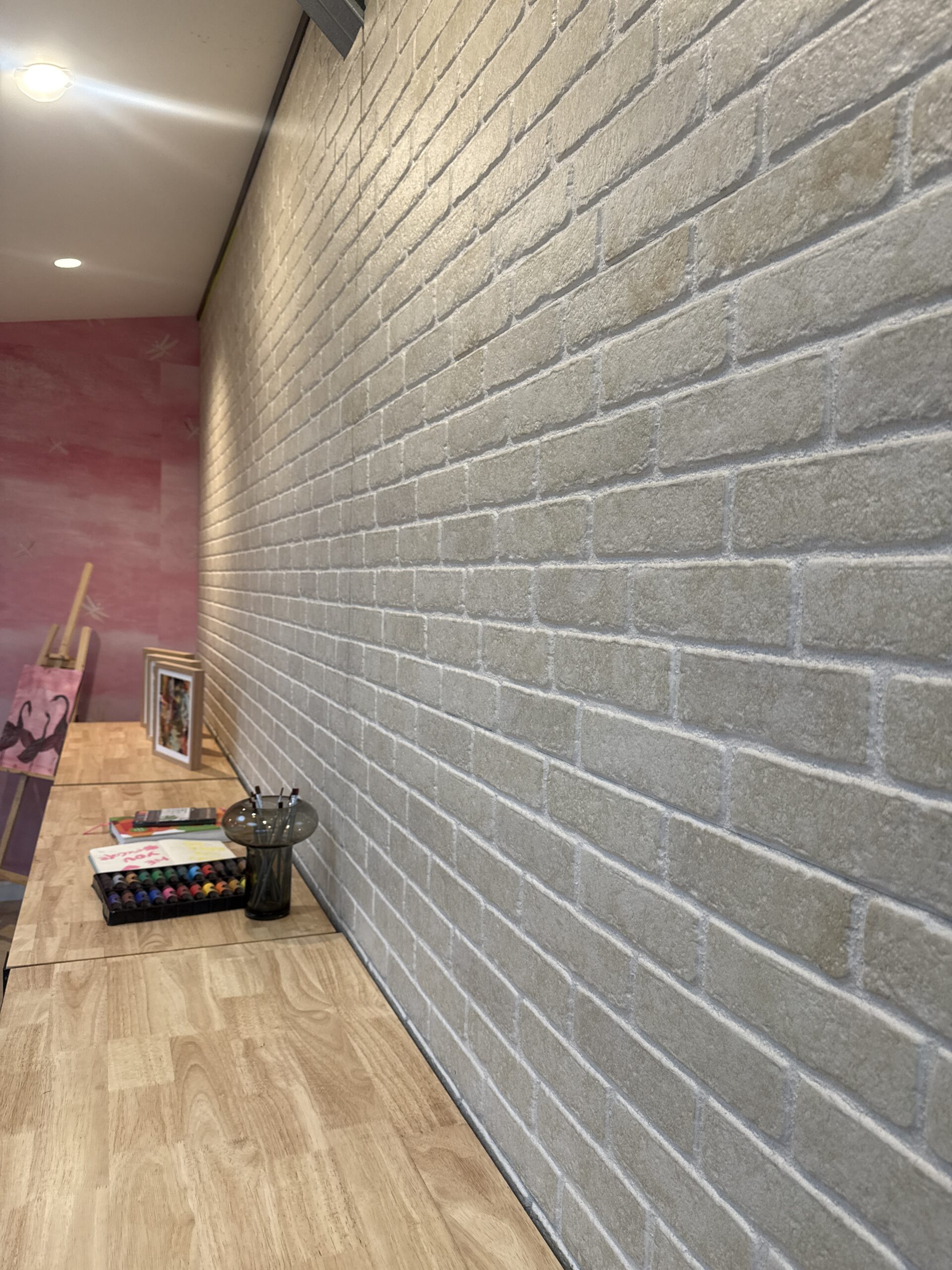 A wooden desk runs alongside a light-colored brick wall. Art supplies, colored pencils, a framed picture, and a painting on an easel are arranged on the desk. The far wall is painted pink.