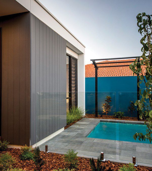 Modern backyard with a small rectangular pool surrounded by gray tiles, landscaped garden beds with mulch and plants, glass fence, and a blue privacy wall with a red-tiled roof visible in the background.
