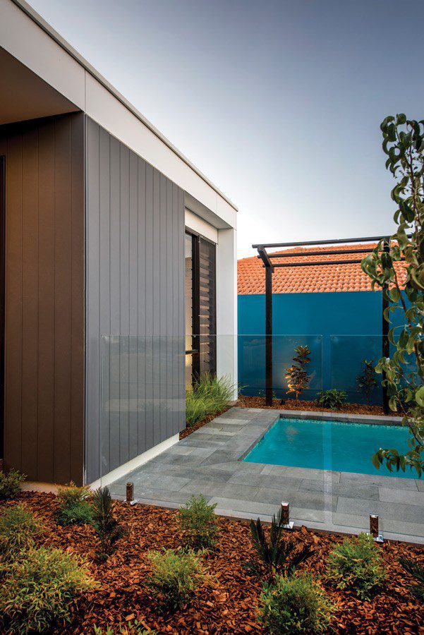 Modern backyard with a small rectangular pool surrounded by gray tiles, landscaped garden beds with mulch and plants, glass fence, and a blue privacy wall with a red-tiled roof visible in the background.