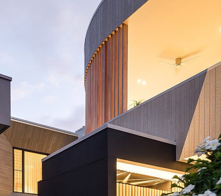 Modern building with curved and angular lines, featuring wood and dark panels, large windows, and warm lighting. White flowers and greenery are visible in the foreground under a cloudy sky.