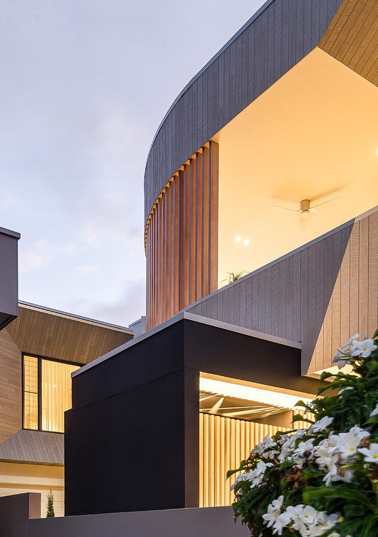 Modern building with curved and angular lines, featuring wood and dark panels, large windows, and warm lighting. White flowers and greenery are visible in the foreground under a cloudy sky.