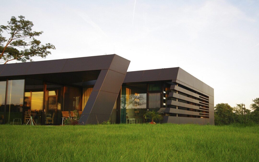 A modern house with sleek, angular black panels and large glass windows sits on a grassy lawn, surrounded by trees under a clear sky.