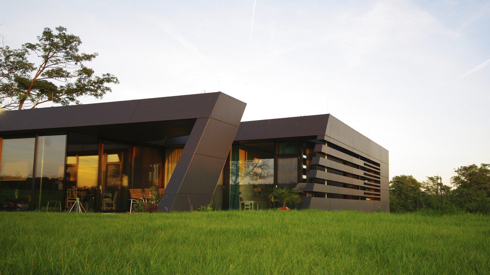 A modern house with sleek, angular black panels and large glass windows sits on a grassy lawn, surrounded by trees under a clear sky.