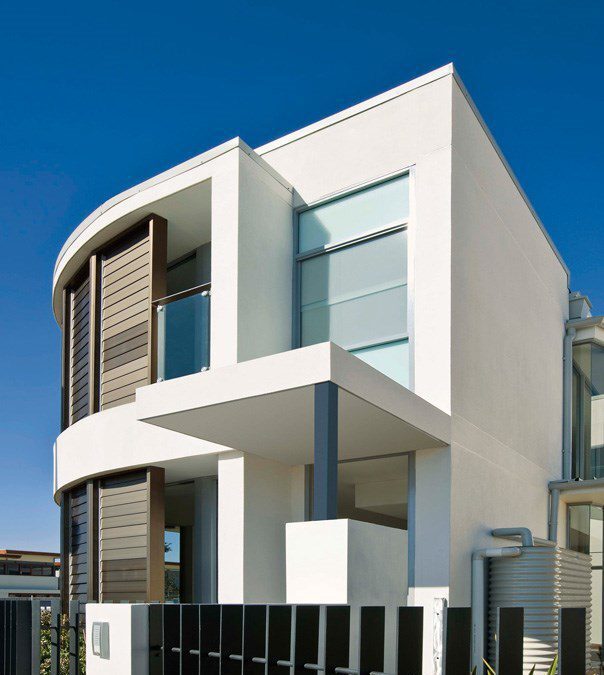 Modern two-story white house with large windows, a flat roof, and dark vertical accents, seen behind a black metal fence, against a clear blue sky.