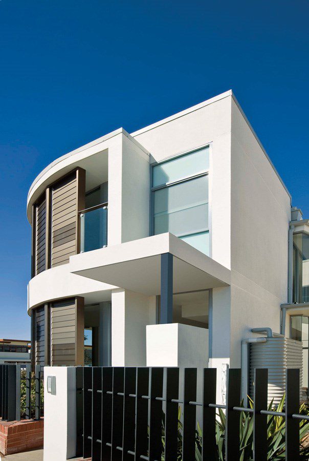 Modern two-story white house with large windows, a flat roof, and dark vertical accents, seen behind a black metal fence, against a clear blue sky.