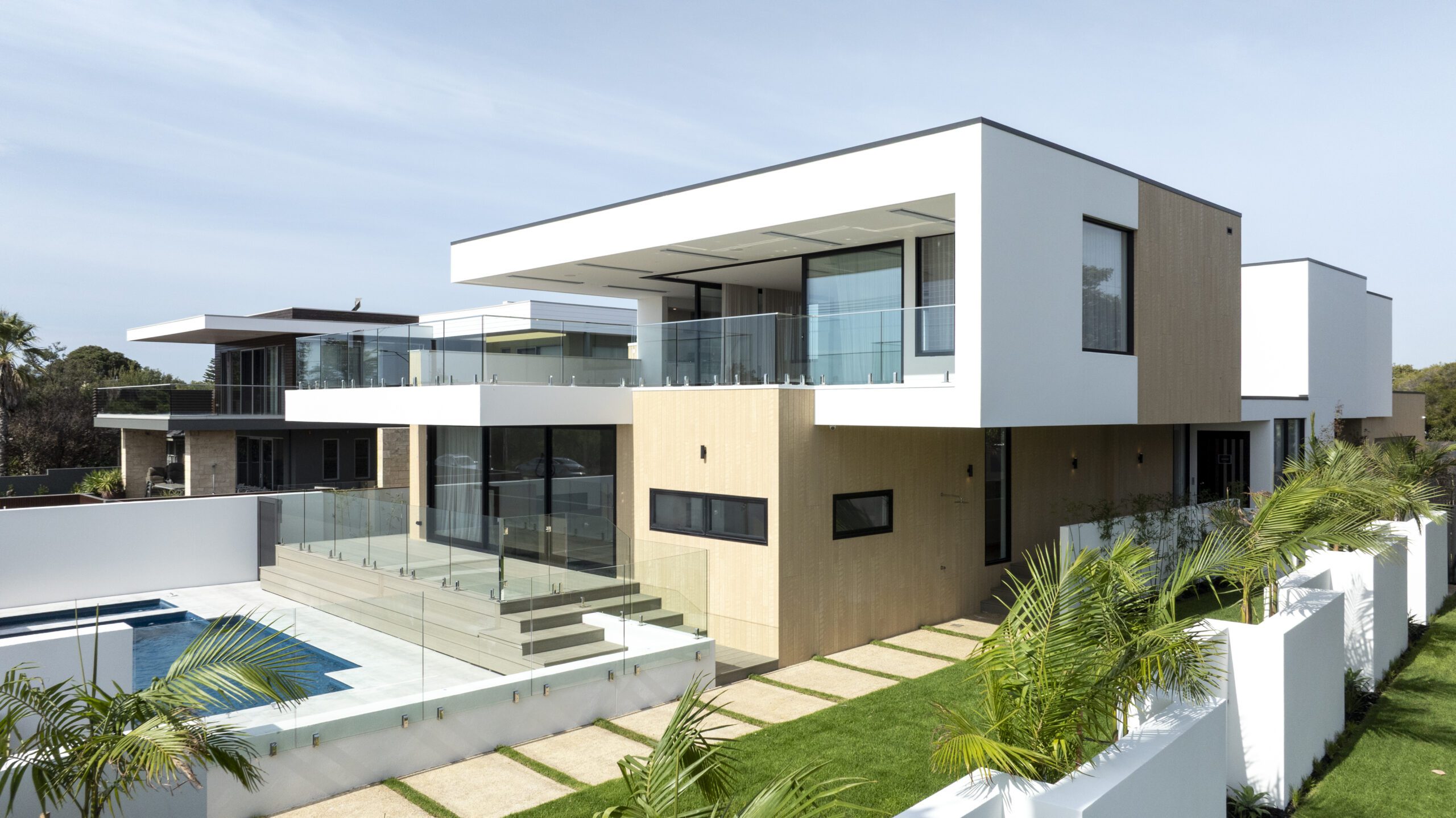 A modern two-story house with large glass windows, wooden paneling, and spacious balconies overlooks a swimming pool. The home is surrounded by a green lawn, palm trees, and a white fence under a clear sky.
